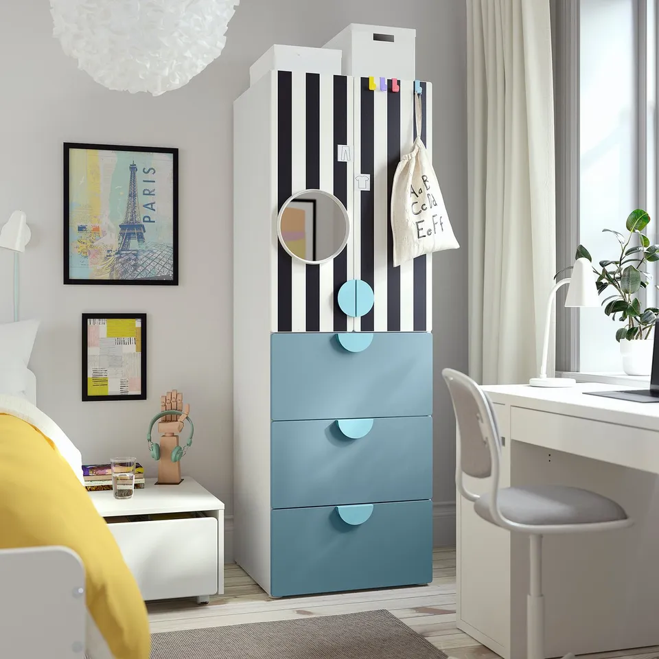 Modern kids room with blue SMÅSTAD wardrobe, striped doors, adjustable feet. Contains black-white art, yellow blanket, white desk & grey chair.