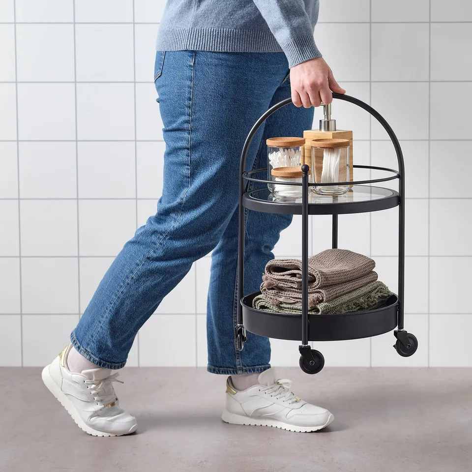 Person pushing a black bathroom trolley filled with folded towels and a wooden container on the bottom shelf, a glass jar on the top shelf.