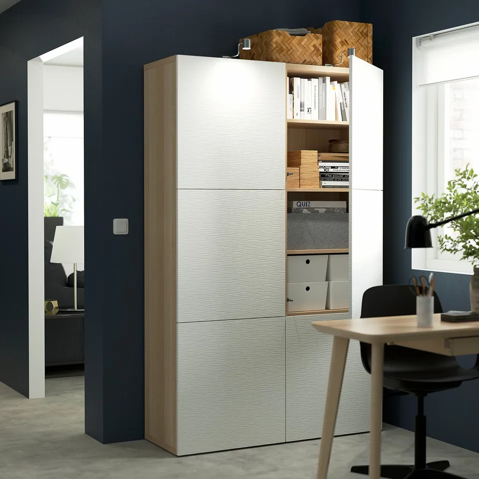 Modern white storage unit, open to show adjustable shelves, books, and filing folders. Adjacent black office chair and wooden desk with a white lamp.