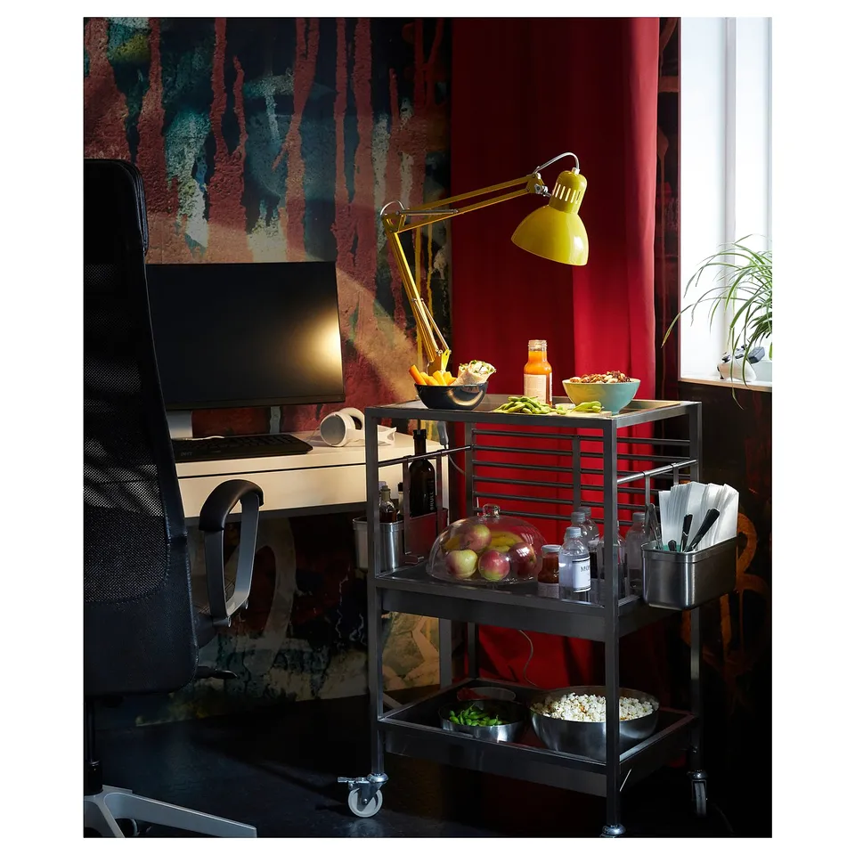 Three-tiered silver metal kitchen trolley under yellow lamp. Holds fruits, snacks, and bottles.