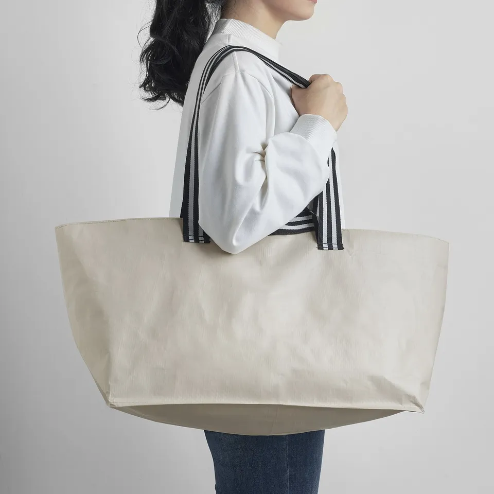 A person holding a large, beige tote bag with black and white striped straps. The bag appears spacious and easy to carry.