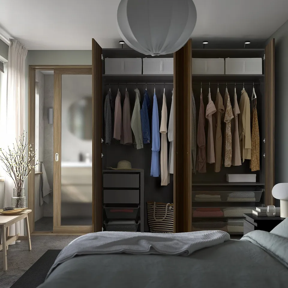 Room with open wardrobe, clothes, drawers, and storage. Light bed, side lamp, large mirror.