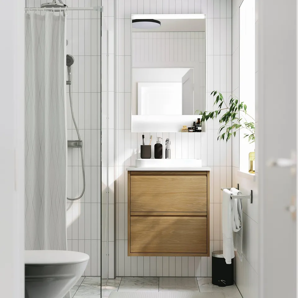 Modern bathroom with light wood vanity, marble sink, glass shower, plant by window.