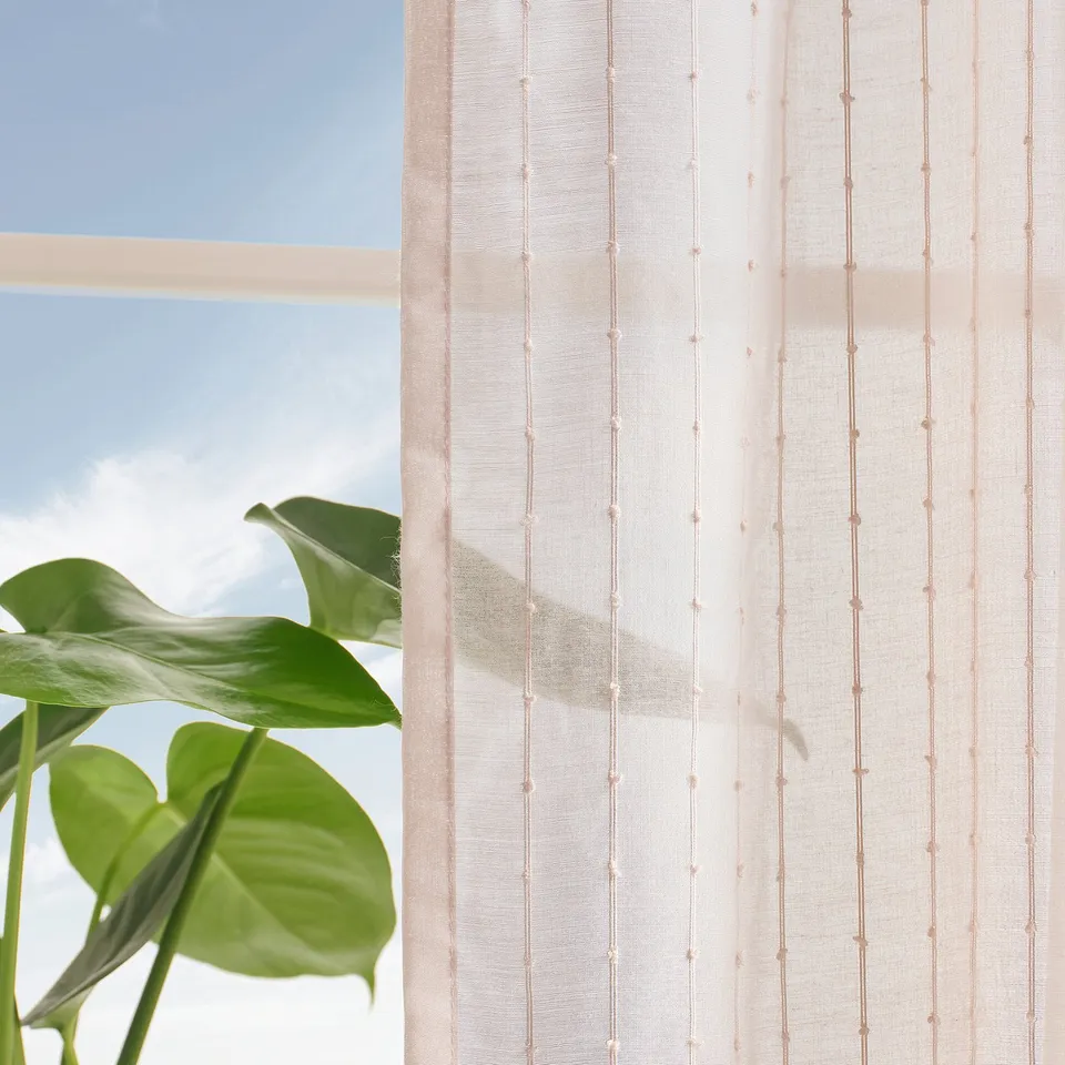 Sheer curtains with pale pink stripes hang by a window, filtering sunlight. Green plant leaves are visible in the foreground.