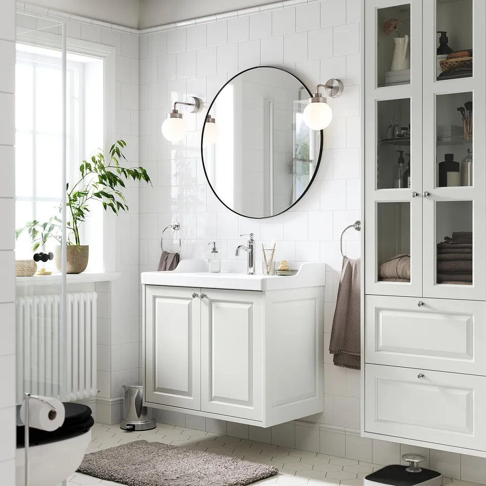 Modern white bathroom with TÄNNFORSEN vanity, round mirror, & wall lights. Plants & towels decorate, providing storage & style.