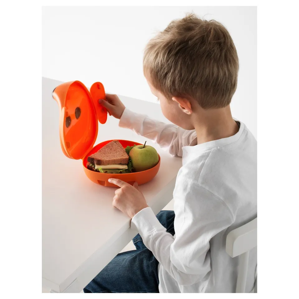 Child sitting, opening orange lunchbox with ears, containing sandwich and apple.