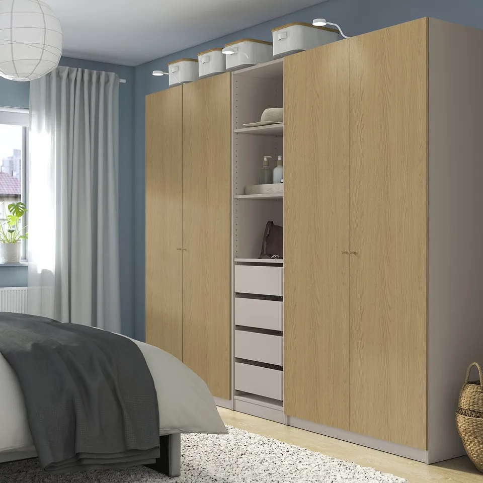 A light wooden PAX wardrobe, tall and spacious, with multiple compartments and drawers. It stands next to a bed with grey curtains.