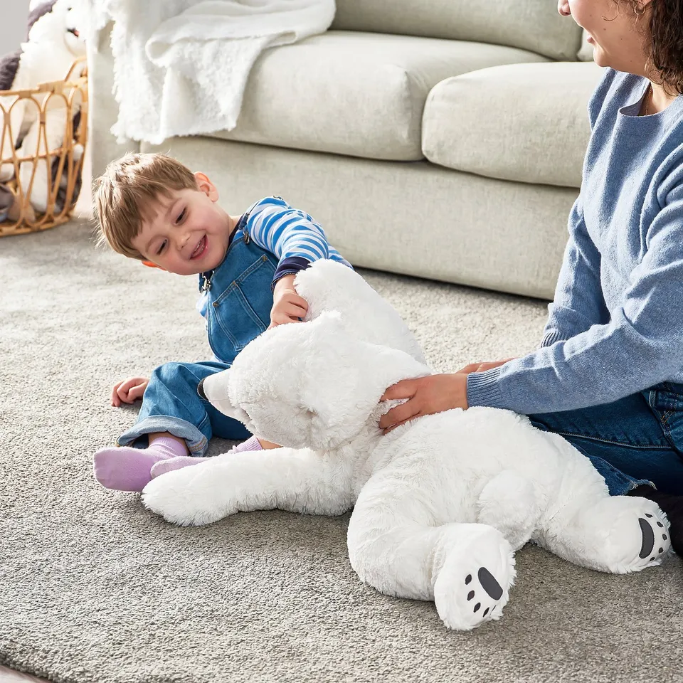 Child playing with large white polar bear toy on the floor. Mother sitting beside them.