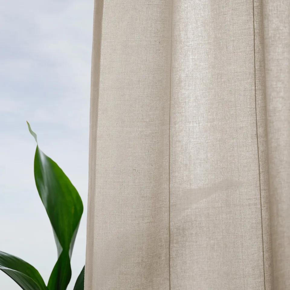 Off-white curtains hang, letting soft light in, with subtle lines adding texture. A plant leaf peeks from the left, showcasing a touch of nature.