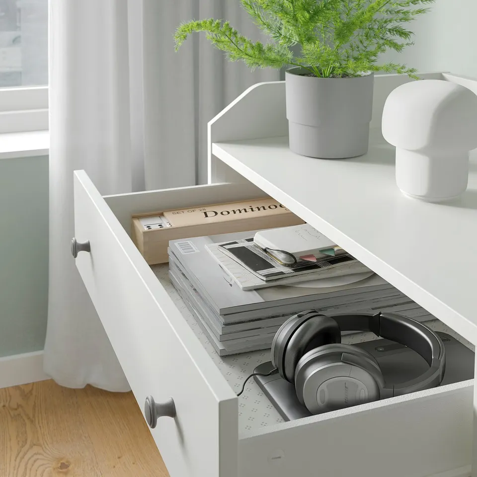 White HAUGA chest with open drawer showing headphones, books, and stationery. Includes potted plant and lamp on top.