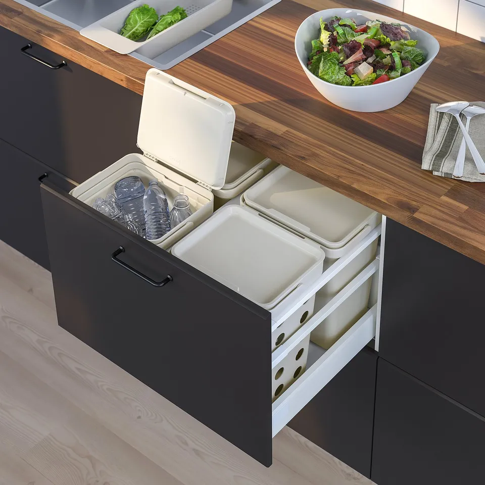 Modern kitchen with built-in waste sorting bins. Dark cabinetry and wooden countertop. Bowls with salad and vegetables nearby.