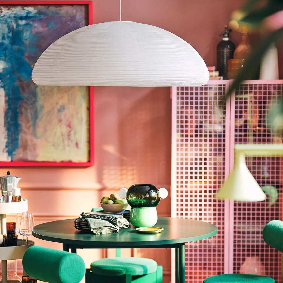 IKEA PS 2026 lamp with white shade illuminates a table with teal chairs, against a pink-tiled wall with art, creating a stylish ambiance.