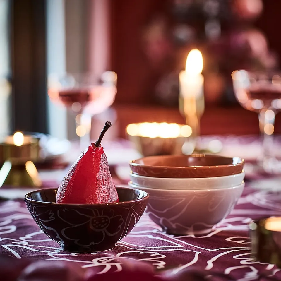 Dark red pear in small patterned bowl on purple floral tablecloth, beside stacked white and brown bowls.