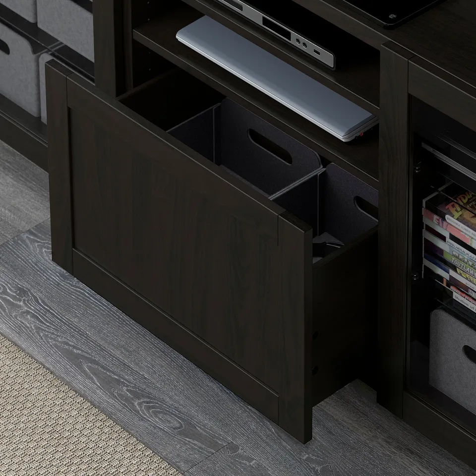 Dark wood BESTÅ storage unit with open shelf holding laptop, closed drawers below, and bins with books beside.