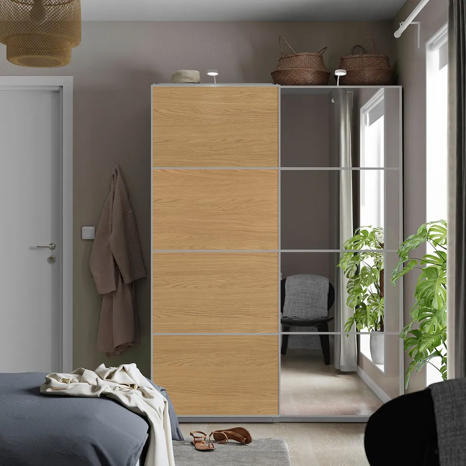 Modern bedroom with light wood wardrobe, mirror finish, white door, grey bedding, baskets, and plant.