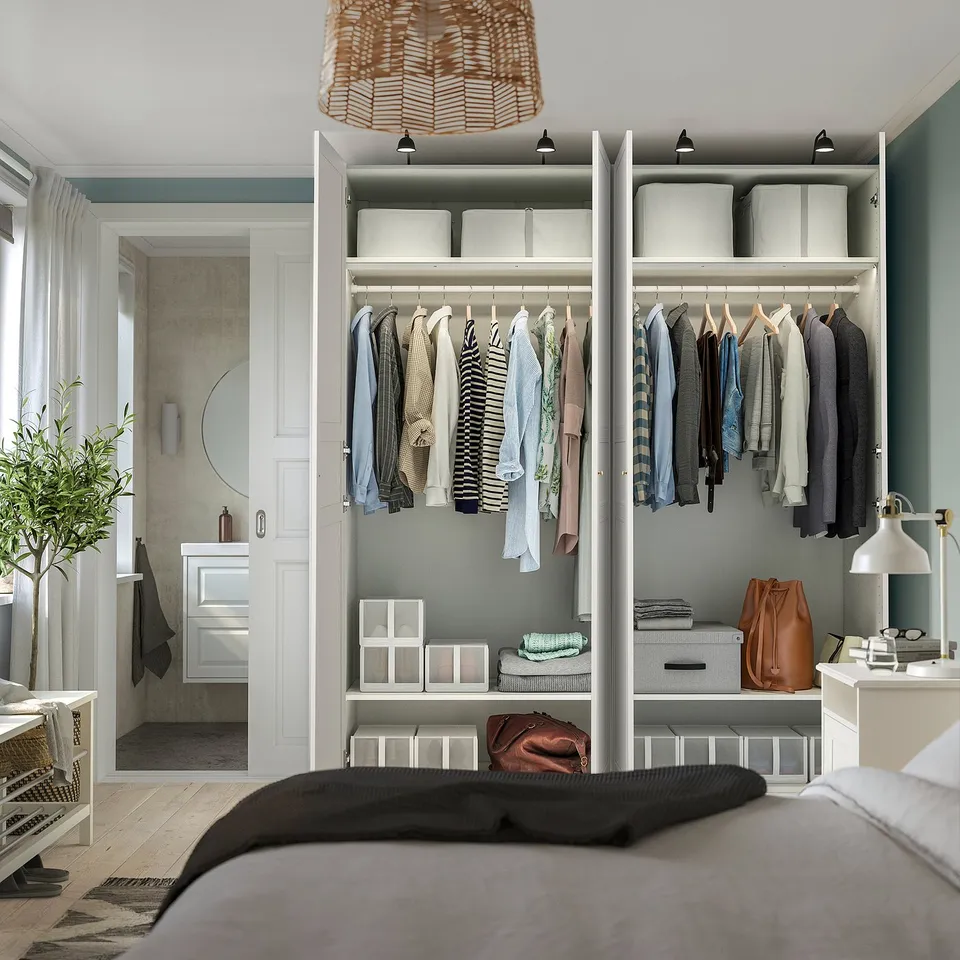 Bedroom with a PAX wardrobe. Wardrobe has clothes hanging, shelves with storage boxes, and a small desk with a lamp.