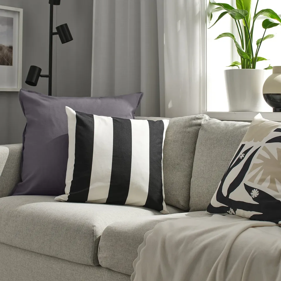 Grey couch with GURLI cushions in solid grey and black/white stripes, recycled cotton. Plant and lamp in background.