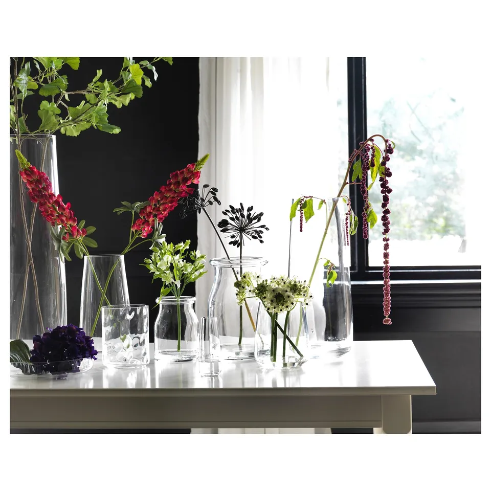 Various vases hold colourful flowers on a windowsill, displaying red, purple, and green blooms, creating a vibrant indoor garden scene.