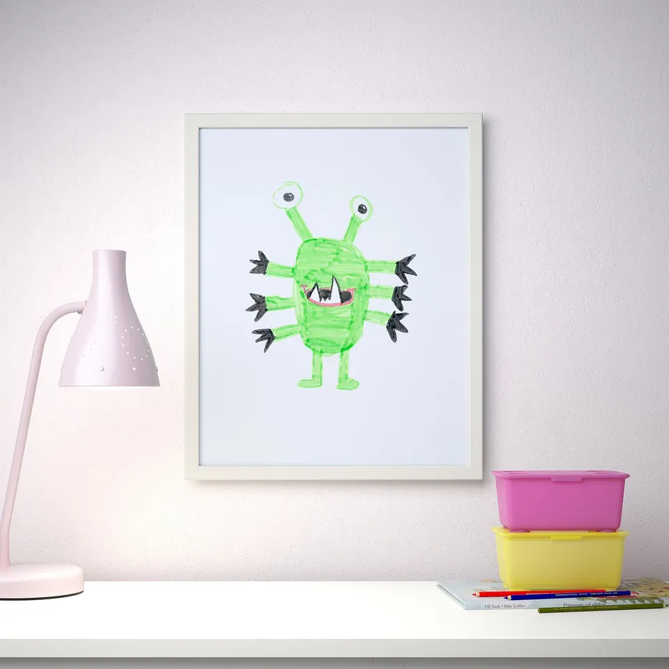 A white desk holds a framed childs drawing of a green, friendly monster. The lamp and storage bins enhance a playful bedroom setting.