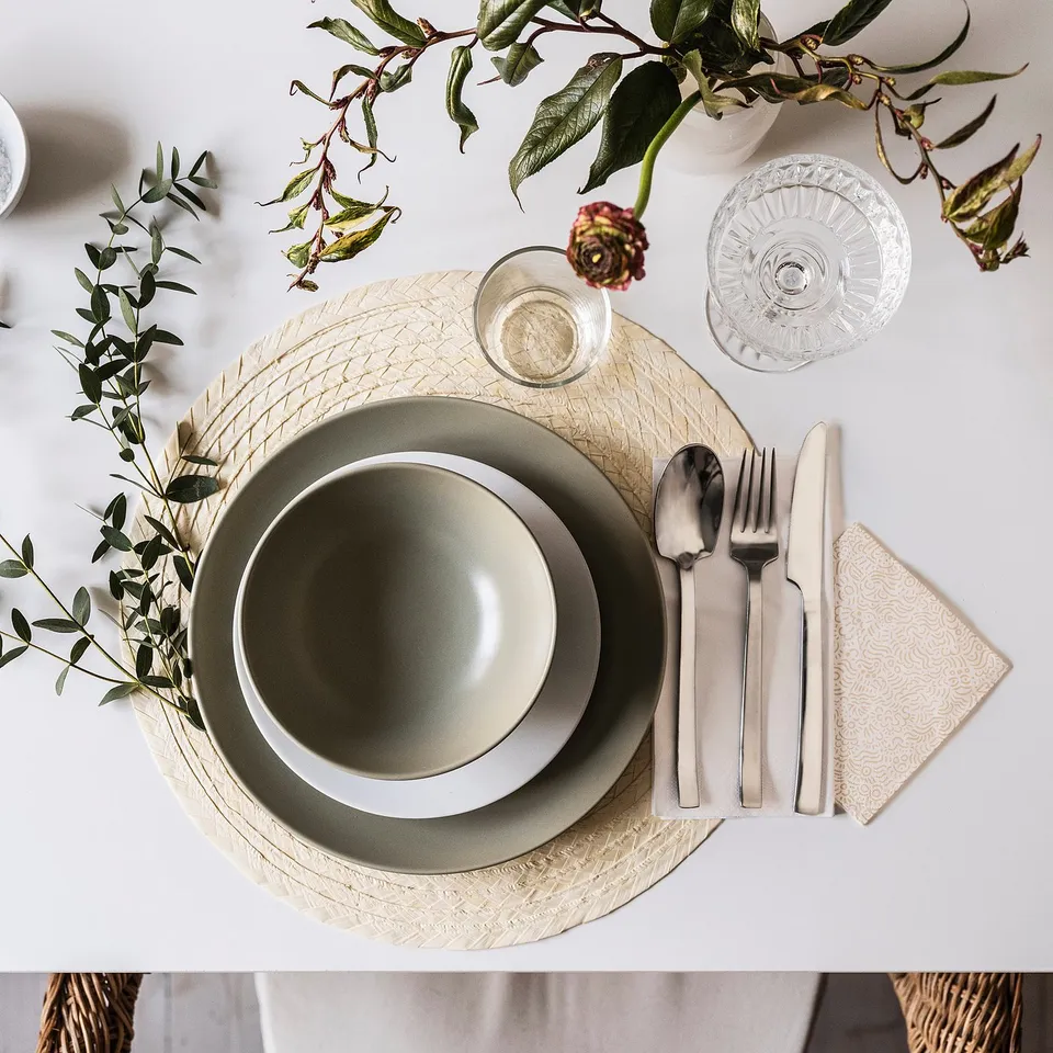 Simple table setting with green plates, metal cutlery, glass, and leafy decor on textured placemat.