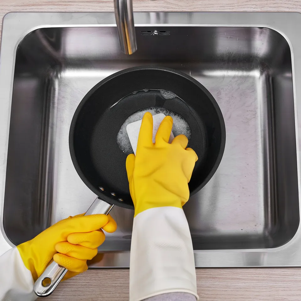 A person wearing yellow dish gloves is cleaning a frying pan in a stainless steel sink. The gloves have protrusions for better grip.