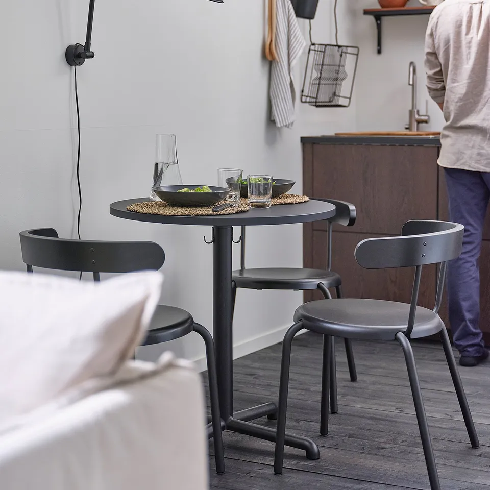 Modern kitchen with black STENSELE table, two chairs, wall lamp, cups, and utensils. Light wood floor and white walls.