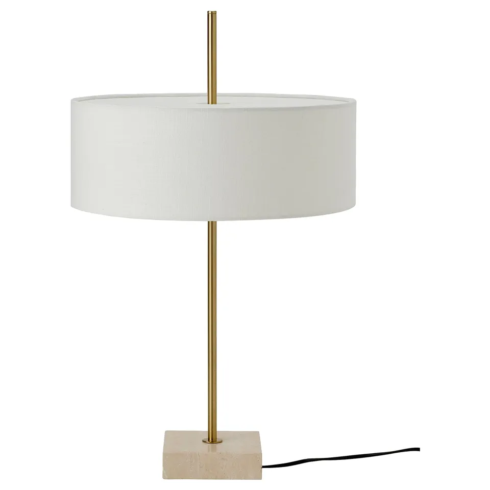 The STOCKHOLM 2025 lamp, showcasing a brass-colored foot, travertine base, and textured textile shade, exuding an elegant design.