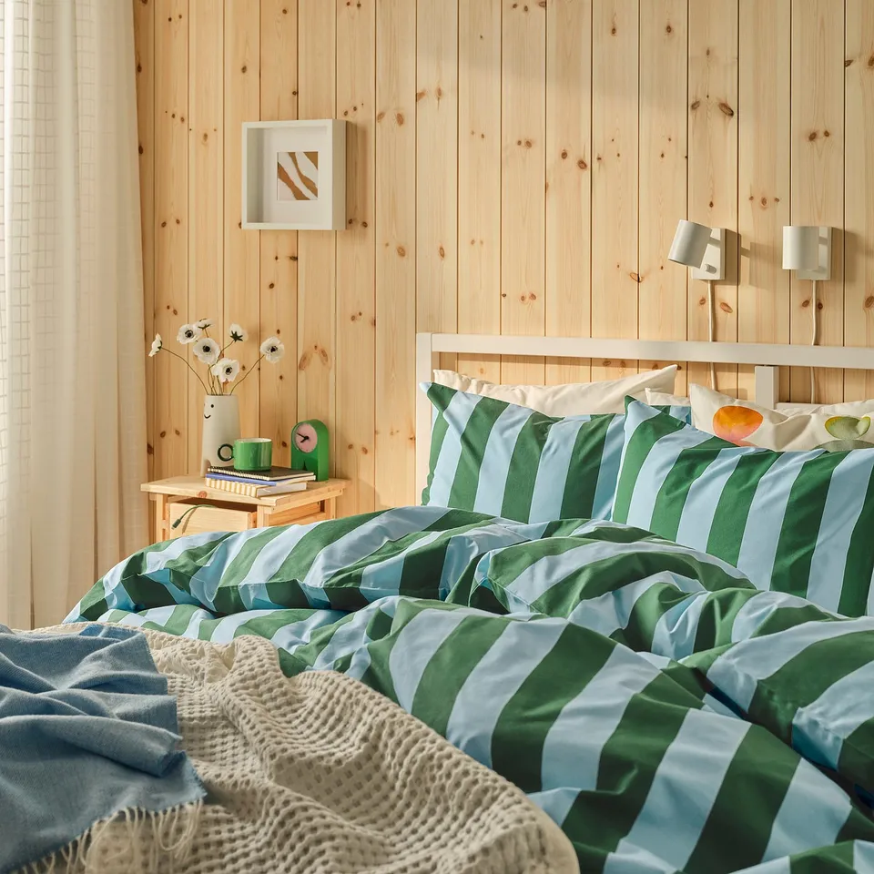 Green-blue striped bed with knitted blanket, side table flowers, cosy curtains.