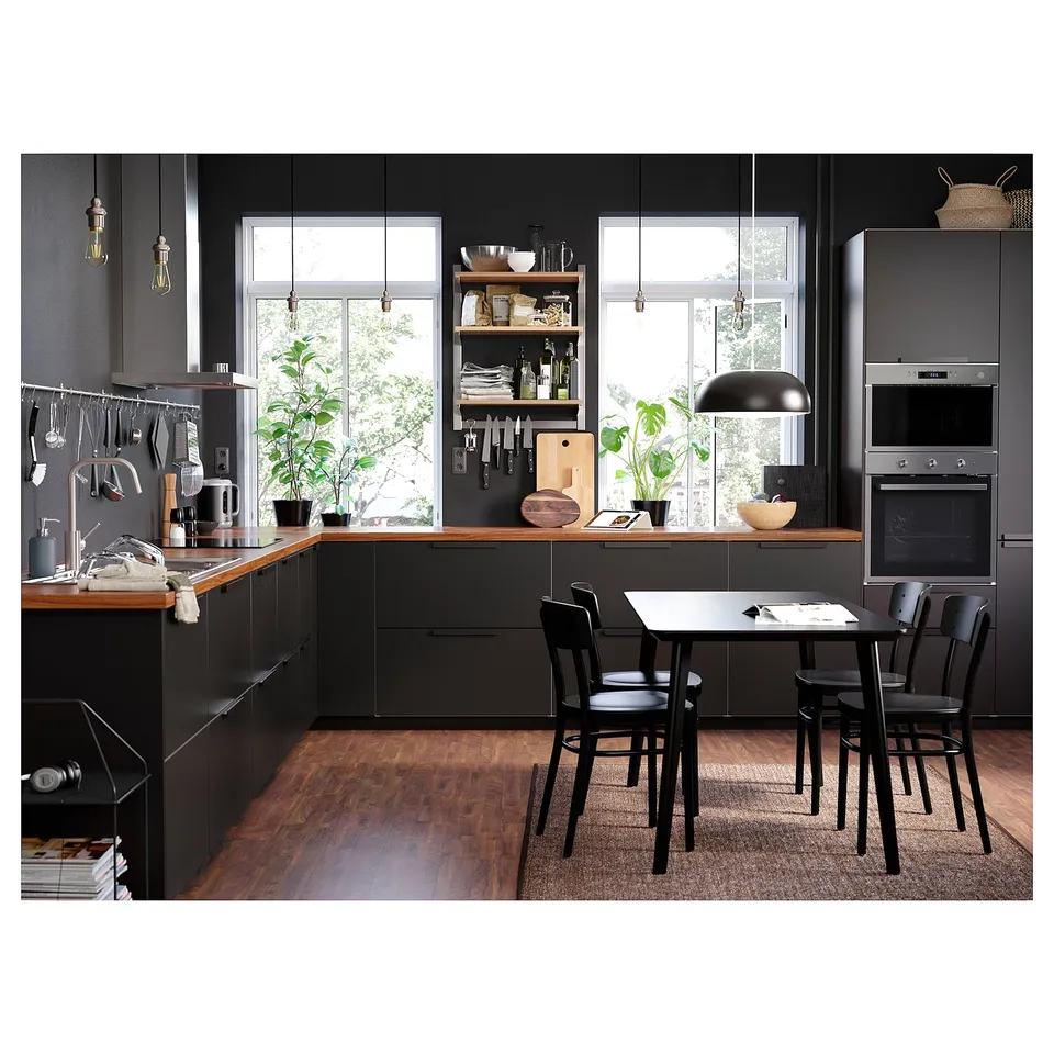 Modern kitchen with black cabinets, wooden counter, hanging lights, plants, and black chairs around a square table near large windows.