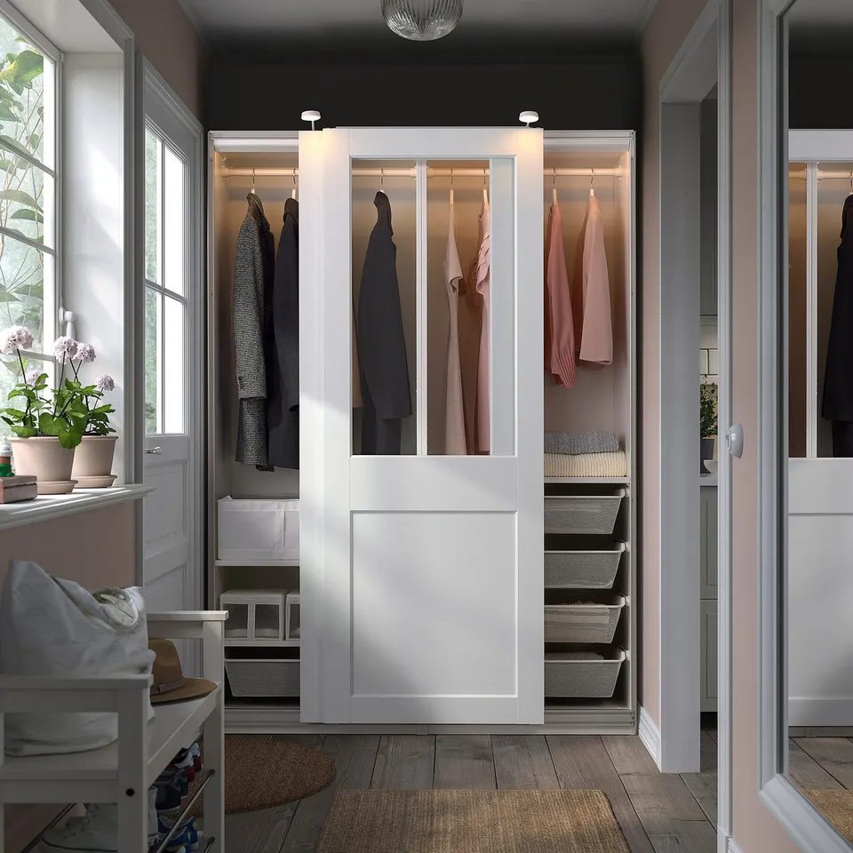 Modern closet with white PAX wardrobes and glass doors, neatly organised clothes and baskets, natural light