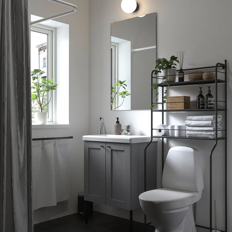 Grey metal shelving unit over toilet, holding items and plants, complementing white fixtures and mirror, with natural light from window.