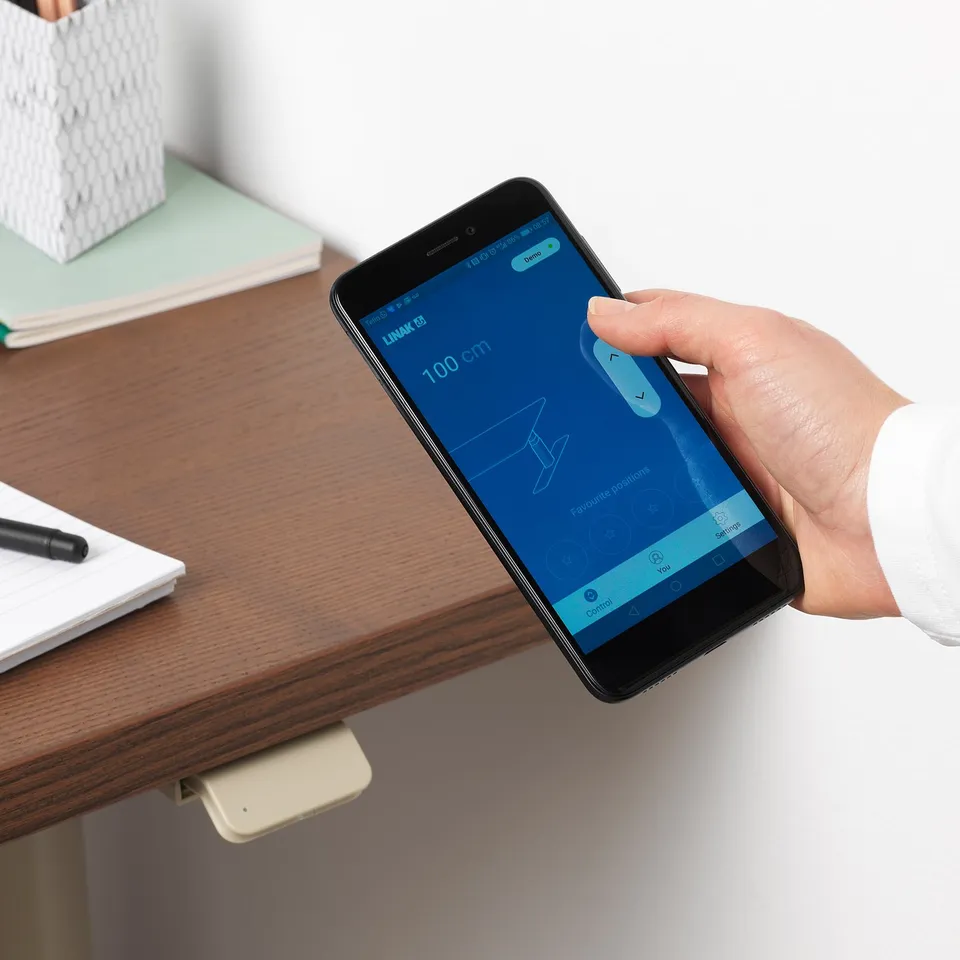 Persons hand holding smartphone displaying IDÅSEN app, adjusting desk height remotely; desk in office setting with notebook and pen.