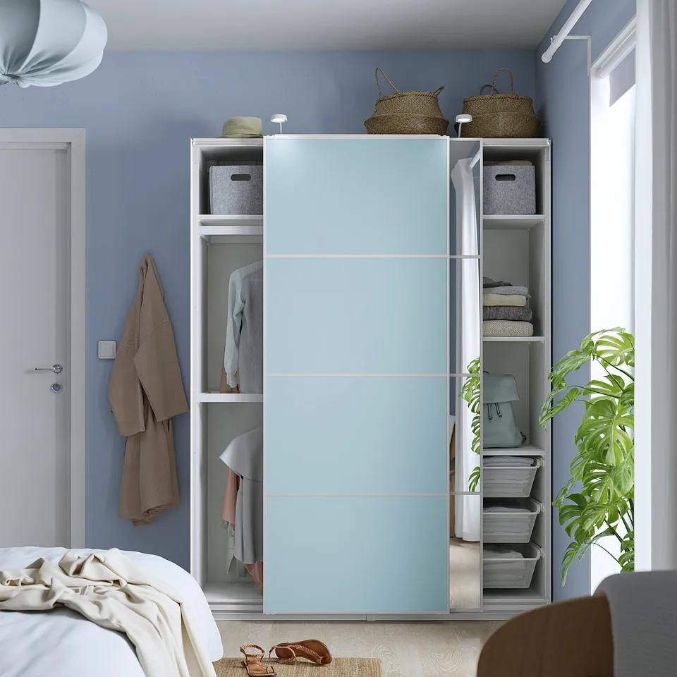 A modern bedroom featuring a light blue PAX wardrobe with sliding doors revealing organised clothes, shelves, and storage baskets.