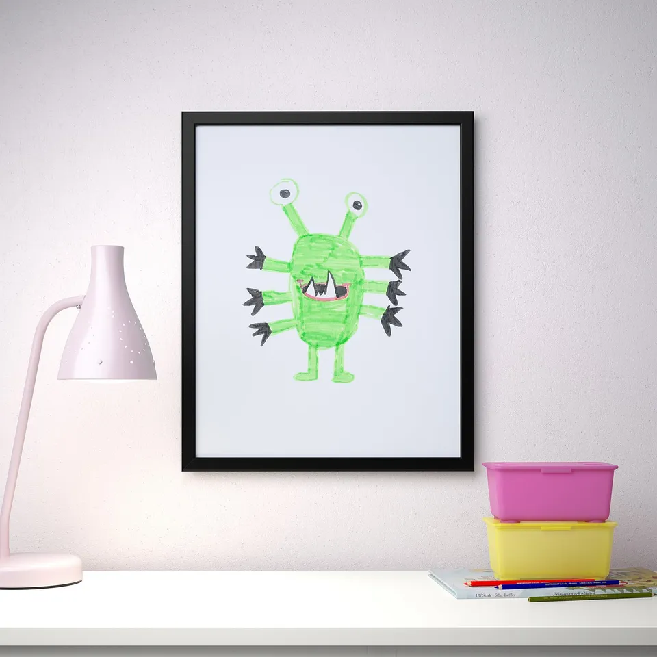A playful monster poster in a black FISKBO picture frame sits on a white shelf. The lamp and storage bins add to the rooms cheerful atmosphere.