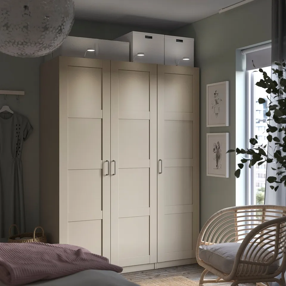 Modern bedroom featuring BERGSBO light beige wardrobe with semi-matte finish, featuring a wicker chair, green walls, and hanging artwork.