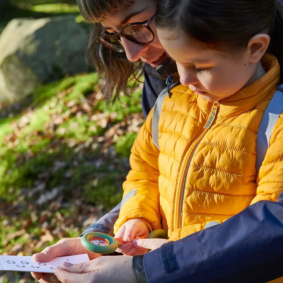 Child and adult use magnifying glass and compass outdoors, examining nature closely.