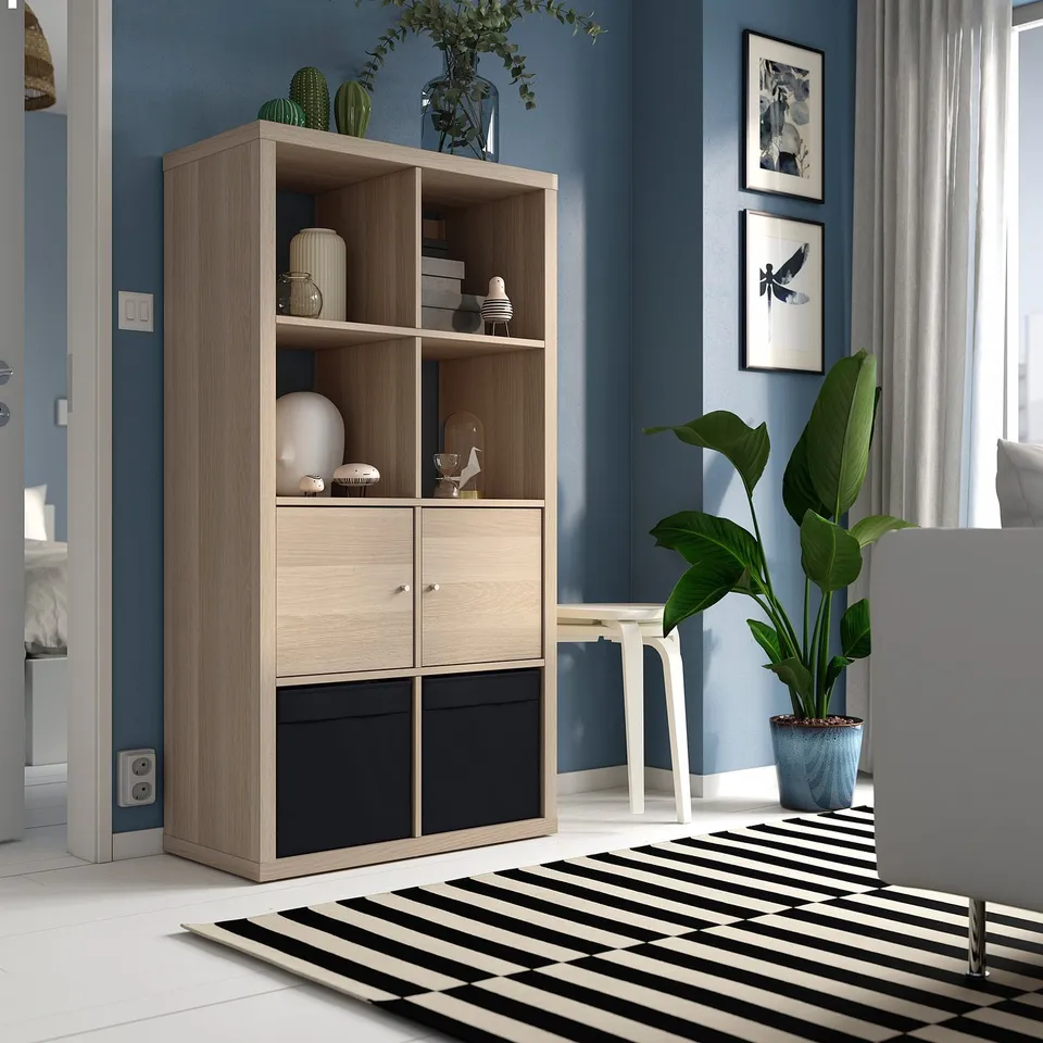 KALLAX light wood shelf with drawers in blue room, holding vases, books, and décor. Surrounded by rug, plant, and chair.