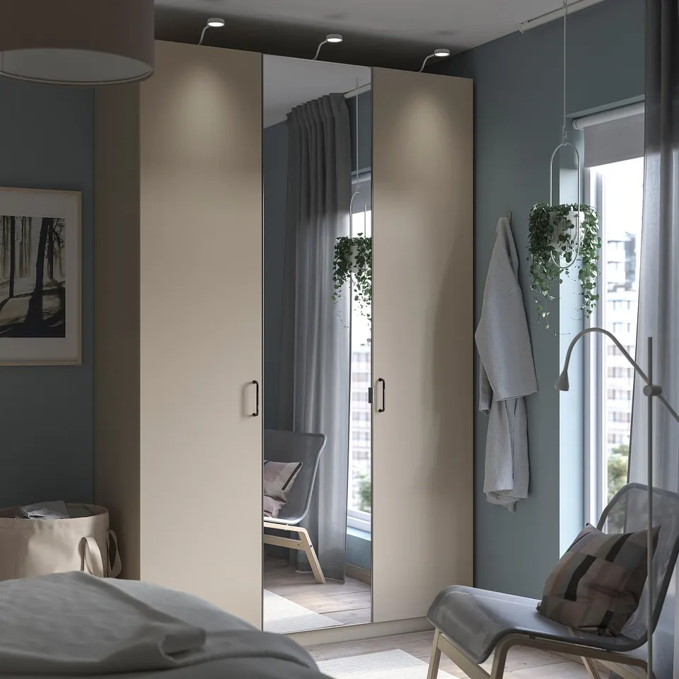A minimalist bedroom a PAX wardrobe with mirrored doors, a hanging plant, and a cosy chair by the window. Grey decor accents the space.