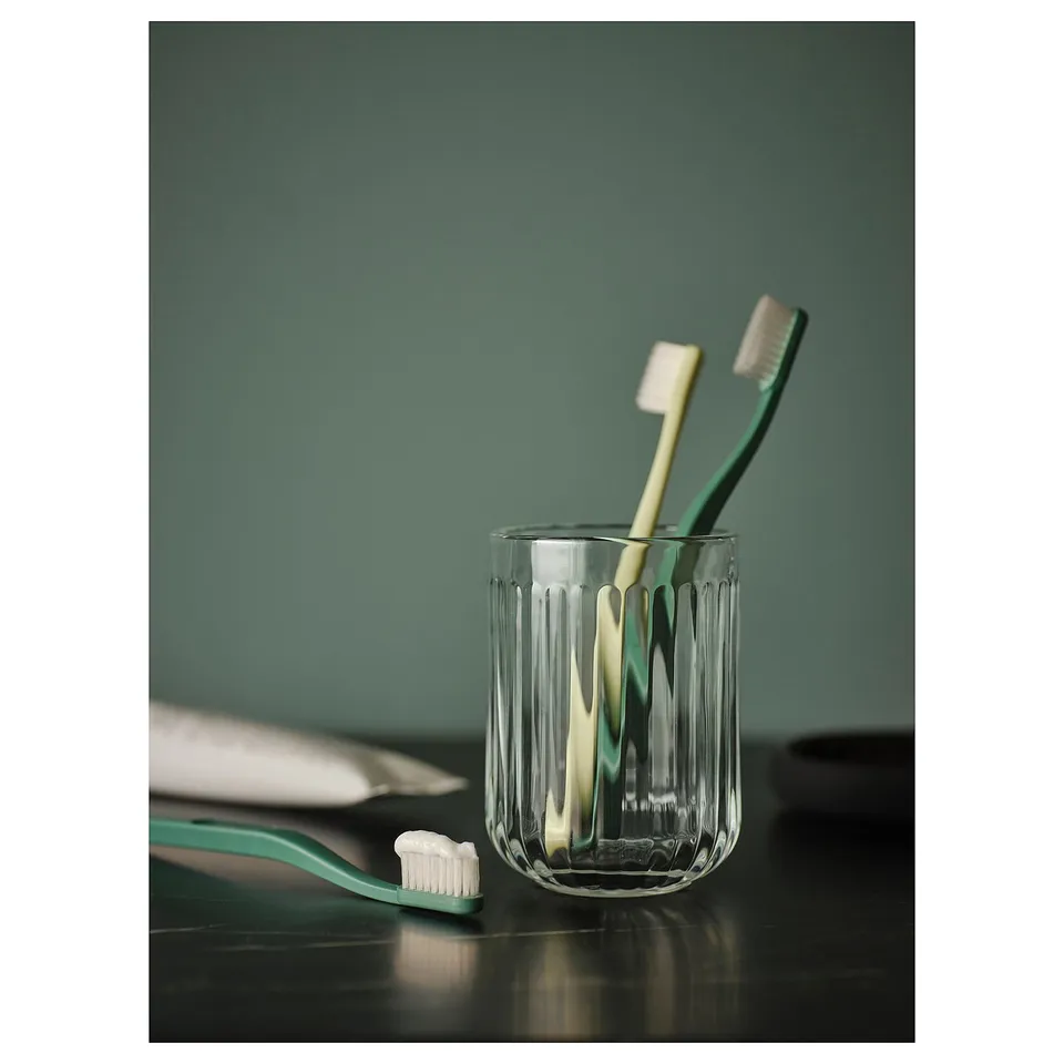 Two toothbrushes in clear glass holder on dark surface. One green, one white. Green wall background.