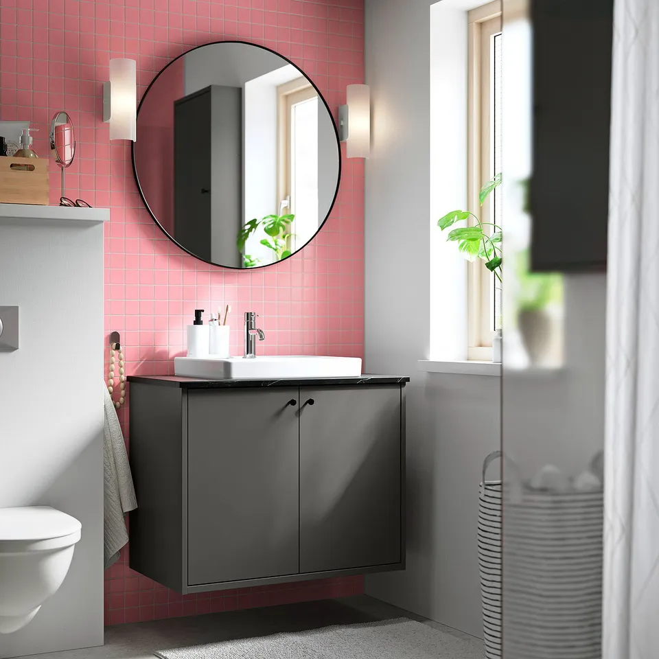 Modern bathroom with grey HAVBÄCK cabinet, round mirror, white sink, pink tiles, and window.