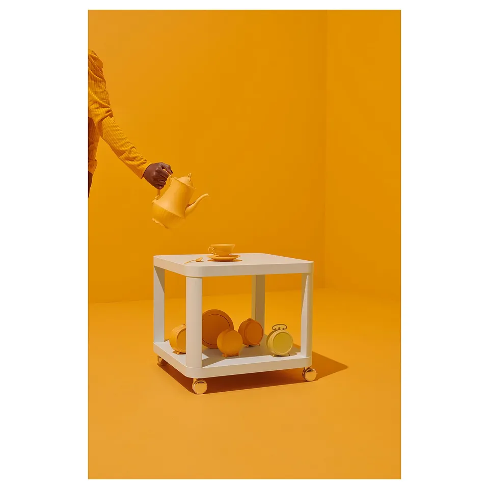 Person holding yellow kettle near white table with yellow items on bright orange background.