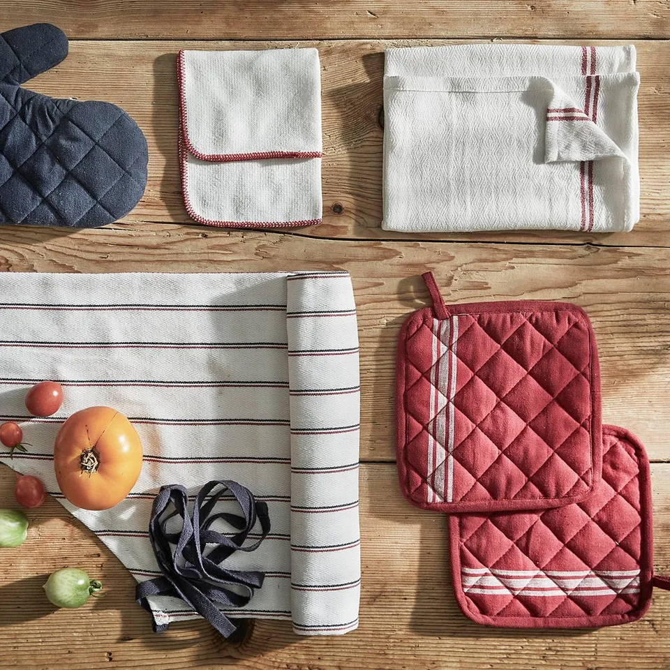 Image shows a set of kitchen linens on wood. Includes striped dish towels, oven mitts, and potholders in blue and red.