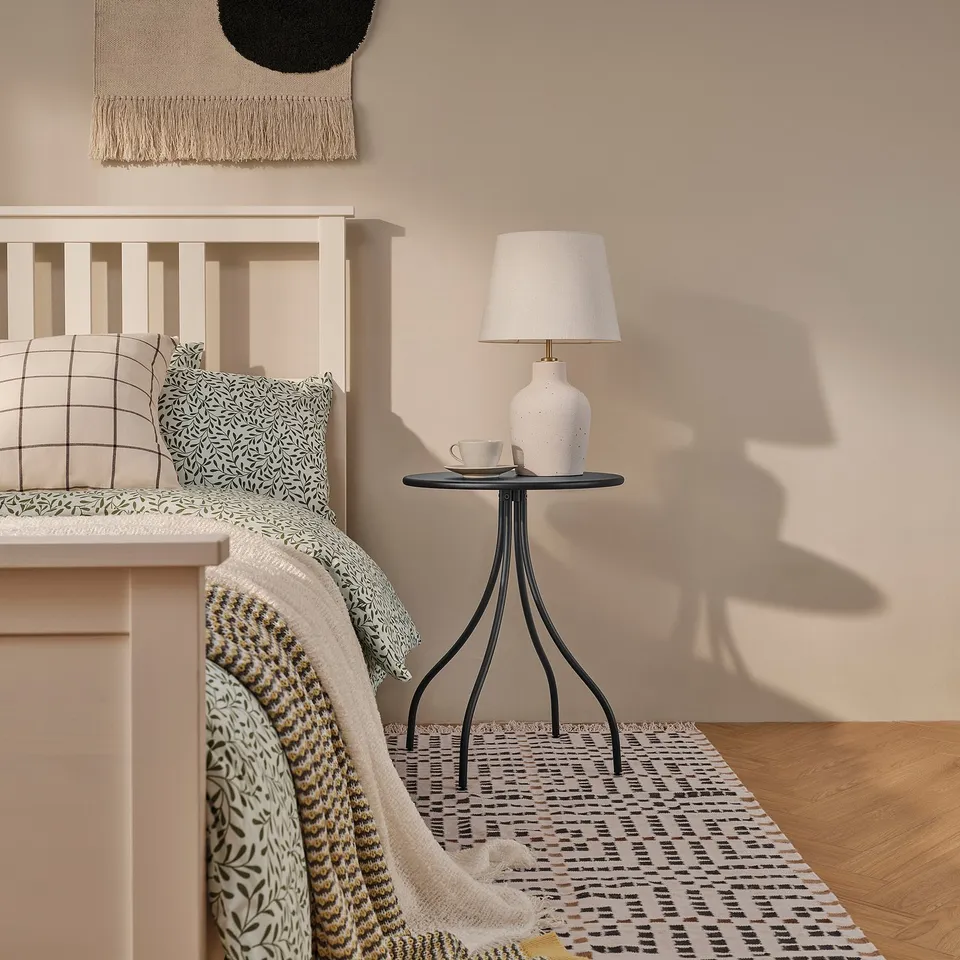 Minimalist bedroom with white bed, patterned pillows, knitted blanket, and TÅNEBRO side table with lamp, cups, and saucer.