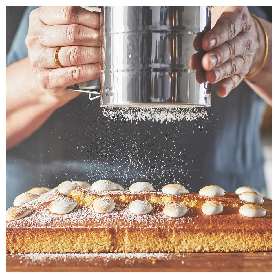 Image shows hands dusting gold cake with icing sugar using silver sifter.