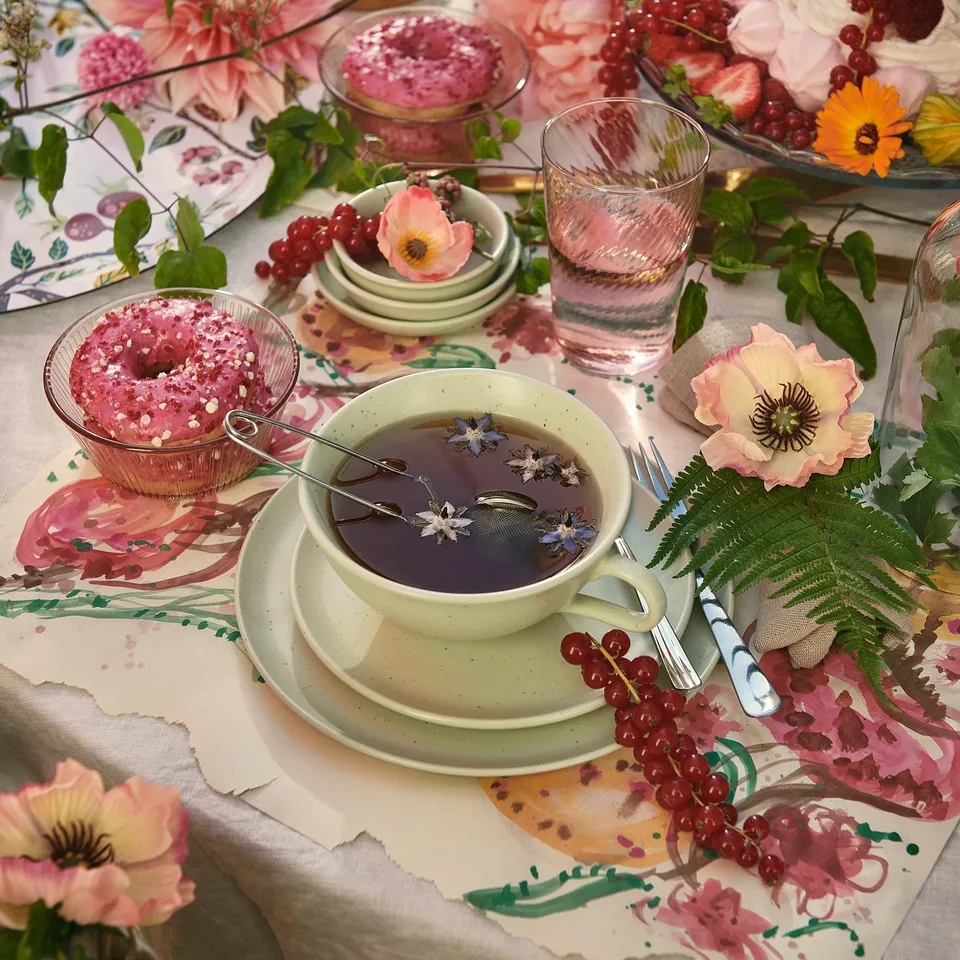 Elegant table set with green mug, glass of water, pink pastries, flowers, and fern leaves on floral tablecloth.