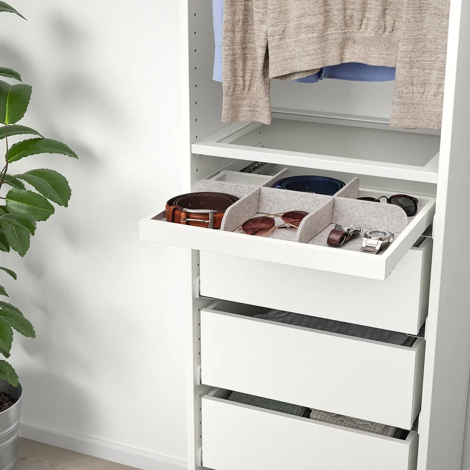 White wardrobe drawer with grey felt trays holding sunglasses, watch, and belt. Plant on left side.