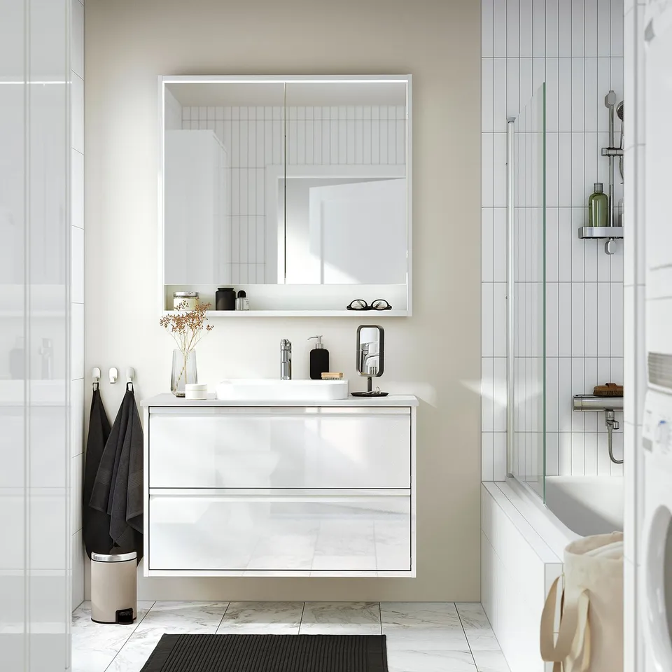 The ÄNGSJÖN vanity, modern, high-gloss white, integrated handles. Its large compared to the nearby shower and bathtub, reflecting minimalist design.