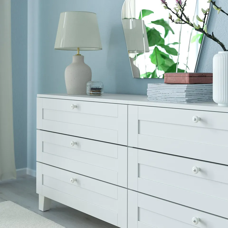White dresser with six drawers, each featuring white porcelain knobs, set against light blue wall. Topped with a lamp and decorative items.