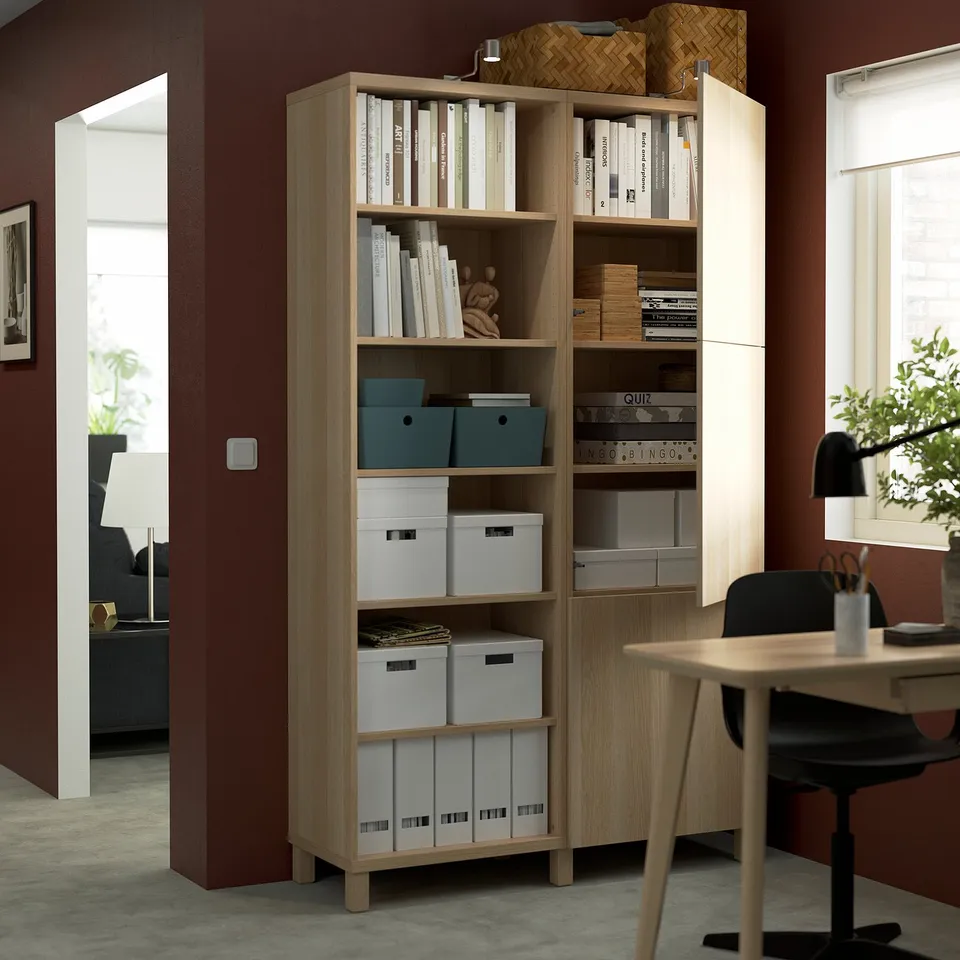 A wooden bookshelf with adjustable shelves storing books, boxes, and decorative items. It is positioned in a home office setting near a desk.