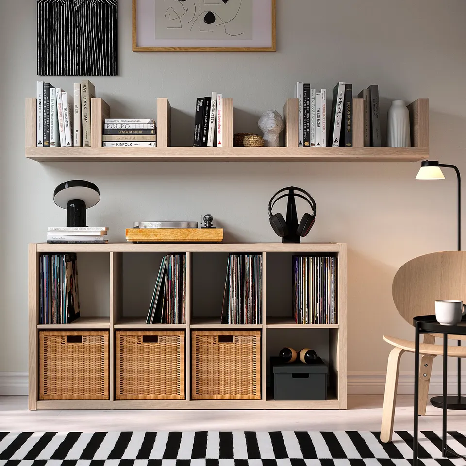 Minimalist living space with LACK shelves, books, albums, decor, bLACK/white rug, wooden chair, side table, lamp, and headphones.